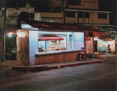 Color photograph of a lit-up taco stand on a street at night.