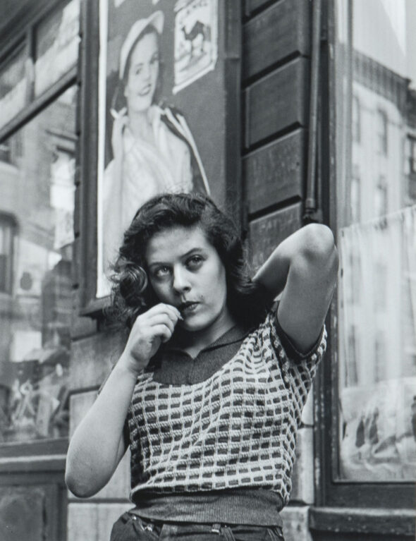 Black and white photograph of a woman leaning against storefront with one elbow in the air and the other pointed down as she puts an object into her mouth.